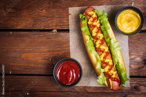 Принти на полотні Hot dog with mustard, ketchup and green salad in bun, served with potato chips on a rustic wooden board