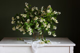 a bouquet of flowering jasmine branches in a glass jar on a white table.