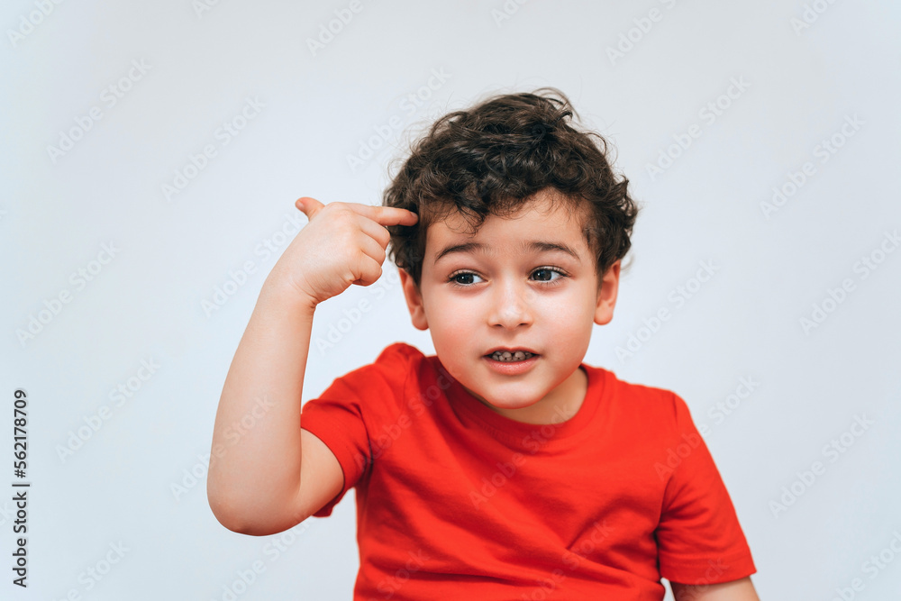 Idea! Smart caucasian baby boy in red t-shirt points at head by index finger with pensive face expression against white wall. Isolated shoot of curly hispanic toddler got a plan. Education, childhood. Idea! Smart caucasian baby boy in red t-shirt points at head by index finger with pensive face expression against white wall. Isolated shoot of curly hispanic toddler got a plan. Education, childhood.