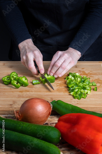 Hands of working woman cutting green pepper on a wooden kitchen board. Decofused gray bowls with eggs, red and green peppers, onion and courgette. Food preparation concept. photo