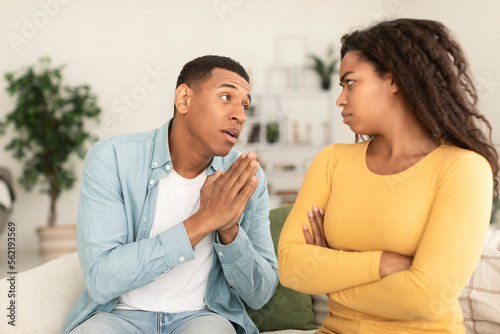 Fototapeta Naklejka Na Ścianę i Meble -  Sad young african american man asking for forgiveness from angry offended female in living room interior