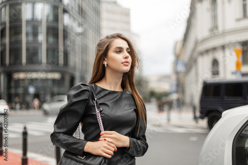 Stylish beautiful business lady in fashion black clothes with a dress and a bag walks in a modern city. Pretty elegant woman