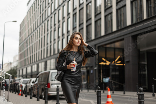 Fashionable beautiful business woman in fashion black clothes with a stylish dress and a leather bag with a cup of coffee walks in the city near a modern office building. Urban pretty girl