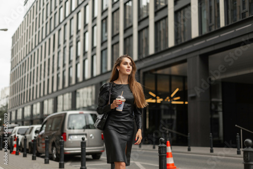 Stylish beautiful urban business woman in fashionable black clothes with a dress and a fashion leather bag walks in the city and drinks coffee near a modern building