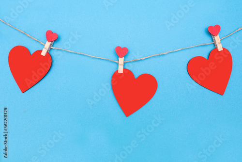 Three red hearts are attached to a rope with clothespins on a blue background. Beautiful decor in the form of hearts or decoration for Valentine's Day. Red wooden hearts on blue paper background