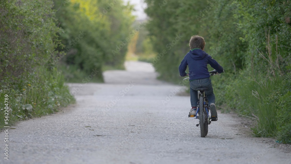 Child riding a bike with back on village road with green trees