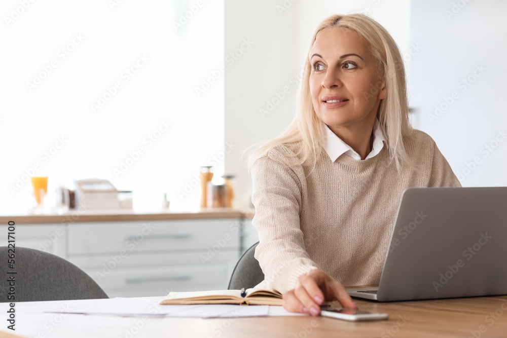 Mature woman with mobile phone and laptop sitting at table in kitchen Mature woman with mobile phone and laptop sitting at table in kitchen