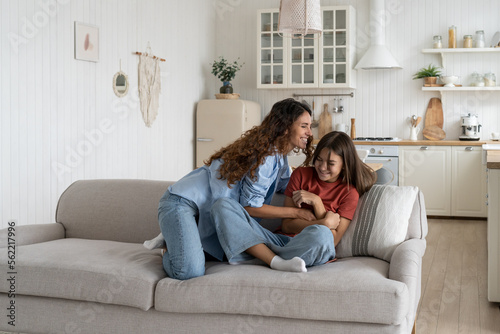 Young European mother tickles daughter to cheer up child lying on couch after hard day at school. Smiling woman and teenage girl dressed in casual clothes enjoy spending time together at cozy home