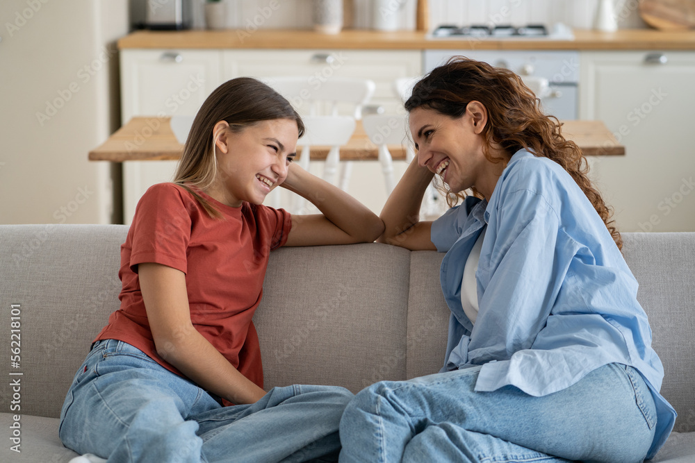 Cheerful friendly family of woman and teenager daughter are sits on sofa and laughing discussing funny school stories. Positive smiling mom and little girl spend time together gossip on weekend 