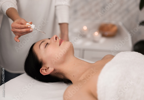Young lady lying closed eyes while cosmetician applying essential oil on face