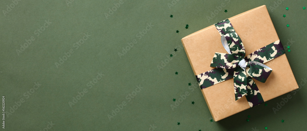 Gift box, tied with a ribbon bow in military colors on green background with copy space. Defender of the Fatherland Day or Veterans Day banner design. Gift box, tied with a ribbon bow in military colors on green background with copy space. Defender of the Fatherland Day or Veterans Day banner design.