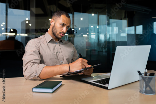 Serious concentrated businessman writing document while sitting in office using laptop for online learning, hispanic man in glasses listening to video call report at workplace.