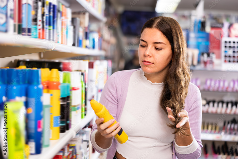 Portrait of positive girl standing near shelves of hair care products and looking at her hair during choosing hairmousse