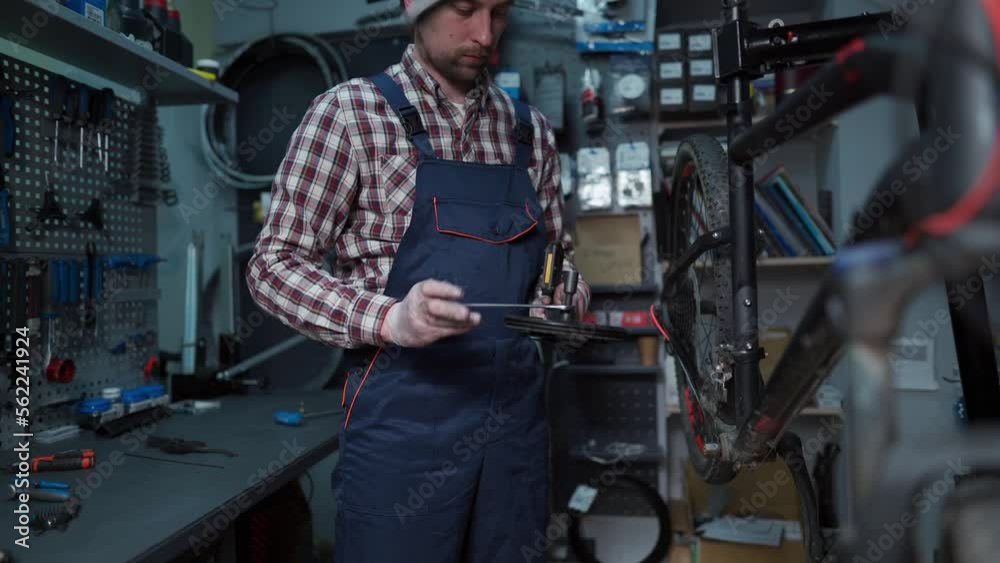 Repairs bicycles in protective clothing at workplace in mechanics repair shop. Bicycle technician adjusts and sets crank on cycle. The crank set and bike carriage. Bicycle renovation profession. Repairs bicycles in protective clothing at workplace in mechanics repair shop. Bicycle technician adjusts and sets crank on cycle. The crank set and bike carriage. Bicycle renovation profession.