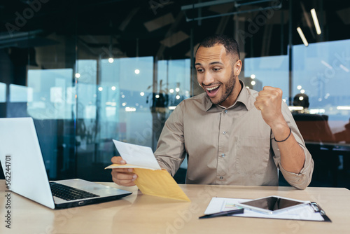 Successful businessman received notification letter with good news, man inside office opens and reads mail envelope with message celebrate victory success, hispanic man holds hand up triumph gesture.