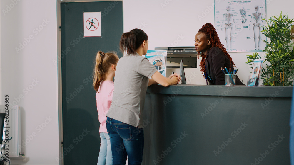 African american receptionist giving assistance to mother and kid, talking about doctor appointment at hospital reception counter. Medical staff filling in checkup report papers in facility lobby. - obrazy, fototapety, plakaty 