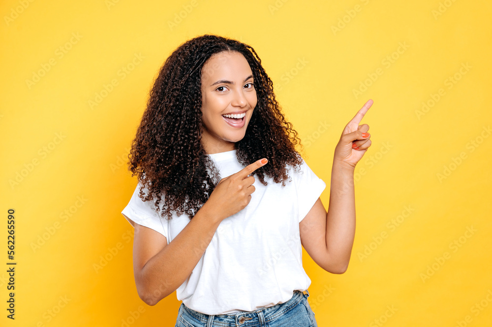 Joyful happy mixed race woman with curly hair, wearing white t-shirt, pointing fingers of both hands to side at empty space, looking at camera, smiling, standing over isolated yellow background