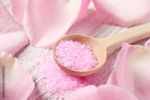 Spoon with pink sea salt and petals of roses on white wooden table, closeup