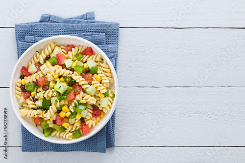 Fresh homemade colorful vegan fusilli pasta salad with beans, corn, tomato, cucumber and green bell pepper, photographed overhead on white wood with copy space on the side (Selective Focus)