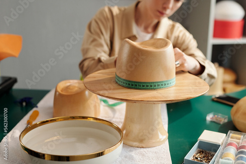 Close-up of wooden workpiece in form of hat against young craftswoman measuring circumference while creating new items for sale