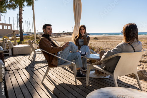 Young friends in warm clothes sitting at table and communicating while spending weekend together in outdoor cafeteria near sea on sunny day