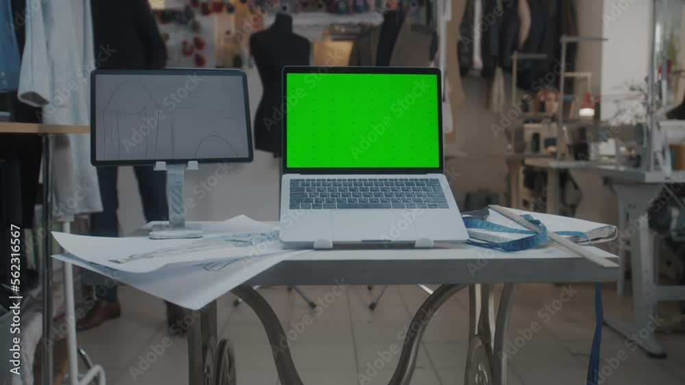 Dolly shot of laptop with chromakey and digital tablet computer with sketch on table in atelier workshop. Tailor and seamstress work on background. Concept of fashion and technologies in business. Dolly shot of laptop with chromakey and digital tablet computer with sketch on table in atelier workshop. Tailor and seamstress work on background. Concept of fashion and technologies in business.
