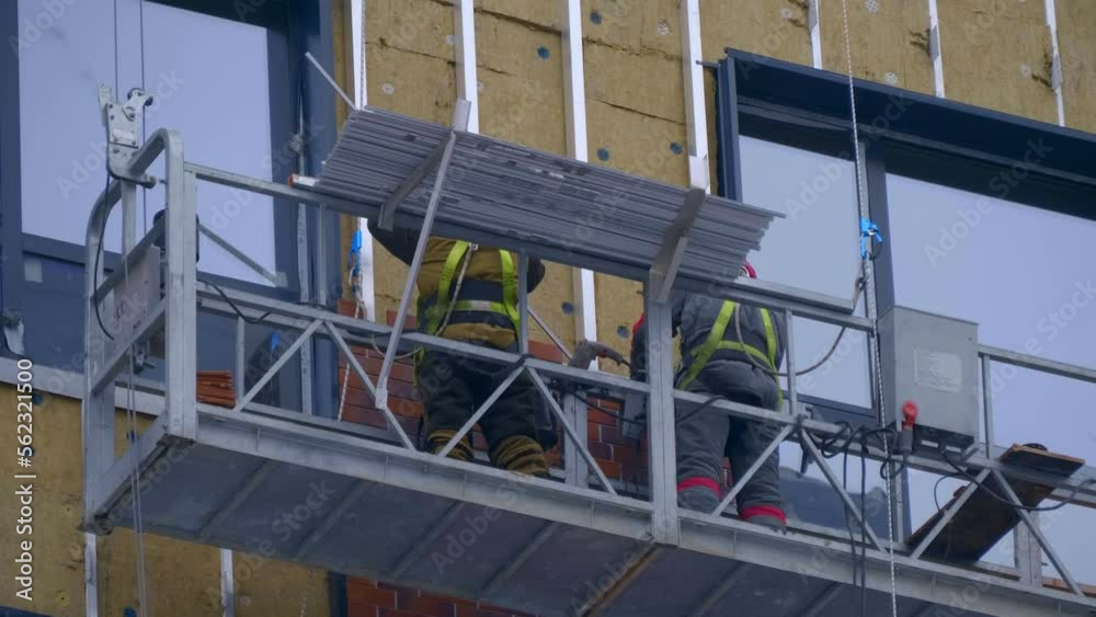 At a construction site, builders in a cradle on cables fastens decorative cladding panels on the wall of a building under construction At a construction site, builders in a cradle on cables fastens decorative cladding panels on the wall of a building under construction