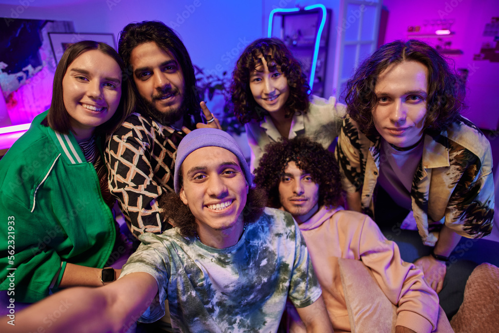 SIx young intercultural men and women posing for selfie in front of camera while enjoying home party in living room lit by neon light SIx young intercultural men and women posing for selfie in front of camera while enjoying home party in living room lit by neon light