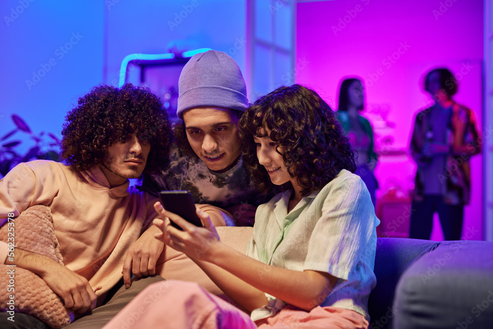 Young pretty woman with dark curly hair and her two intercultural male friends watching online video in smartphone at home party