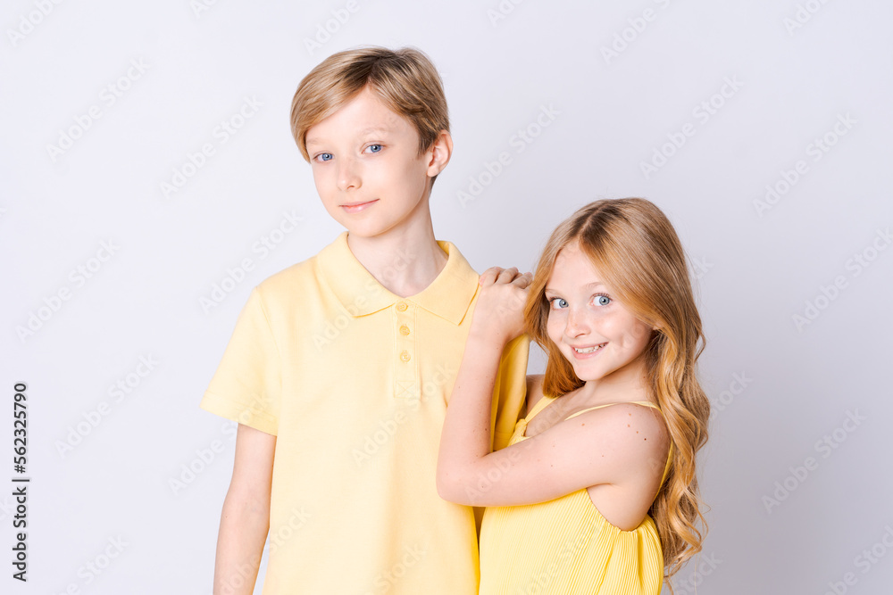 Two happy children standing near an empty gray background and hugging cute little girl with long hair is hugging cute blonde girl showing her love and care Brother and sister have fun posing in studio Two happy children standing near an empty gray background and hugging cute little girl with long hair is hugging cute blonde girl showing her love and care Brother and sister have fun posing in studio