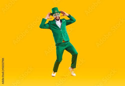 Happy funny guy wearing party outfit having fun on St Patrick's Day. Cheerful joyful positive young man dressed in green suit and leprechaun hat dancing isolated on bright yellow color background