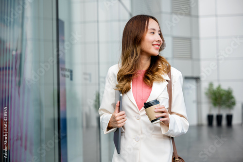 Young business woman wearing white suit jacket and smile while go to work outdoor mirror building background. Confident Businesswoman with cup of coffee holding Laptop walking outside office building © sorapop