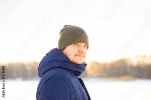 Young man in winter on a snowy lake, portrait. European caucasian white male in hat and down jacket in sun rays.