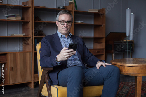 Confident handsome elderly businessman with grey hair in blue suit and glasses working on his mobile phone indoor on office wall with boockshelf
