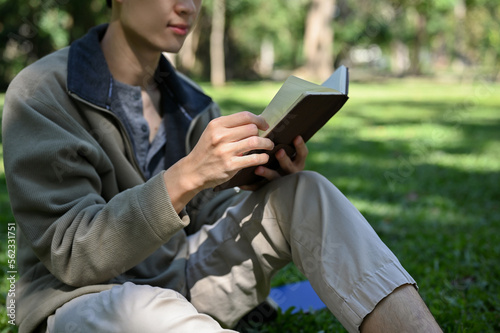 Relaxed Asian male college student chilling in the greenery park on the weekend, reading a book