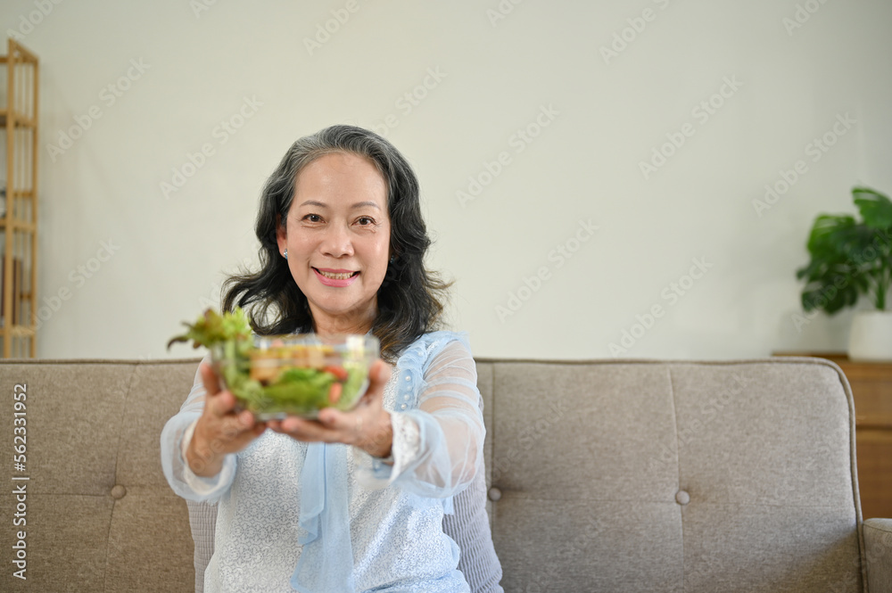 Charming Asian 60s aged woman sits on sofa in her living room and holding a salad bowl. Charming Asian 60s aged woman sits on sofa in her living room and holding a salad bowl.