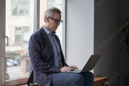Mature businessman in eye glasses and blue suit is working on his laptop on the background of window