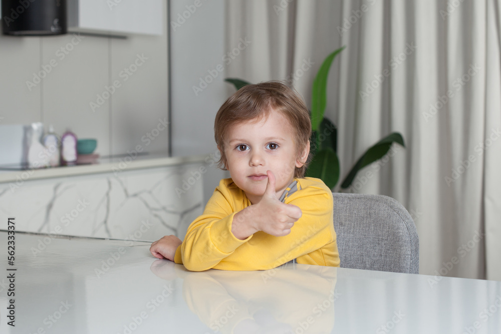 Smiling happy child boy with brown hair in yellow sweater sitting by the table and showing thumb up on studio kitchen background Smiling happy child boy with brown hair in yellow sweater sitting by the table and showing thumb up on studio kitchen background