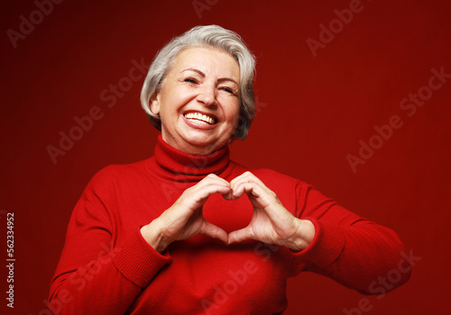 Fototapet Senior grey-haired woman wearing red sweater smiling in love doing heart symbol shape with hands
