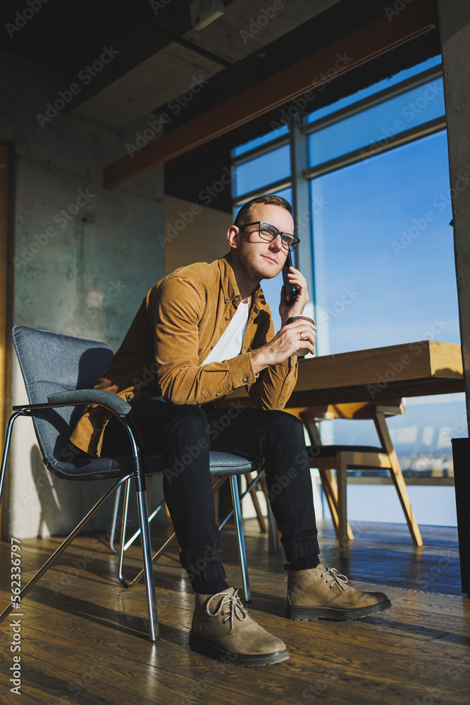 Smiling male worker in casual clothes looking away while talking on mobile phone and drinking coffee during break in modern creative workspace