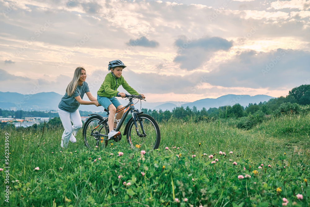 Obraz premium Mother teaching son to ride bicycle. Happy cute boy in helmet learn to riding a bike in park on green meadow in summer day at sunset time. Family weekend. 4K video Slow motion