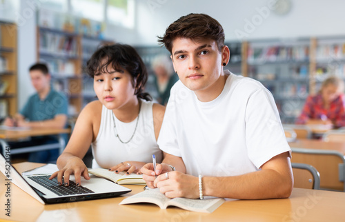 Asian schoolgirl, studying with a classmate guy in the school library on a laptop and making notes in a copybook, preparing for lessons