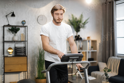 Young attractive sporty man in white t-shirt having morning workout at modern light apartment. Muscular strong guy jogging while training at treadmill.