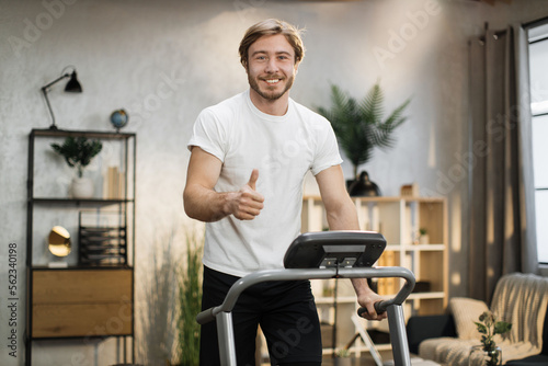 Caucasian muscular sporty man in white t-shirt leaned his hands on treadmill while working out in morning at living room at home, looking at camera showing thumb up.