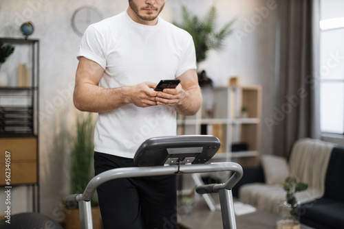 Cropped view of young sports man using phone looking online fitness class while working out, running, doing cardio training on treadmill indoor on background of modern apartment