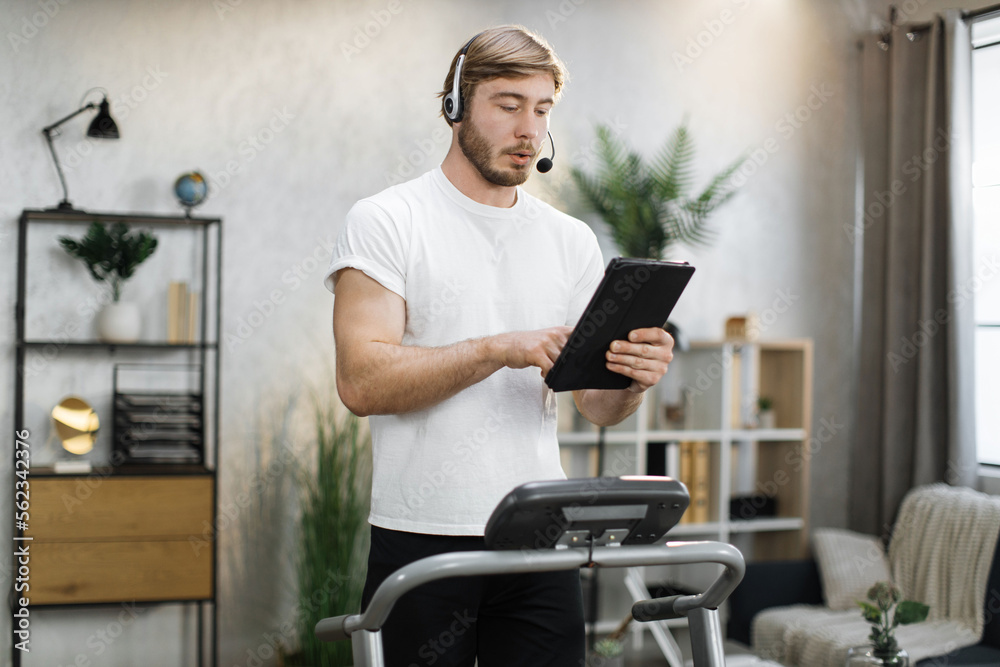 Portrait of active and dynamic young handsome businessman with headset and tablet doing sport fitness at home running on treadmill indoor at night. Tone your body. Perfect shape. Improving endurance.