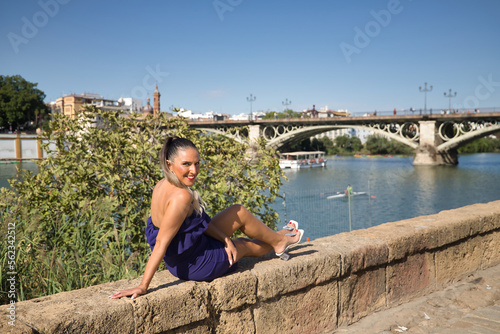 Pretty young woman in a purple suit, sitting on a stone wall by the river, posing happy, Concept beauty, fashion, trend, elegance. © Manuel