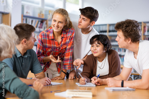 Group of fifteen-year-old schoolchildren are discussing something and making notes in copybooks, preparing for classes in the school library