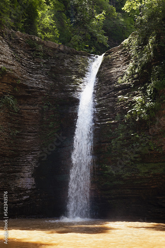 Beautiful hight waterfall in fresh lush green forest in sunny hot  summer day with sunshine and beams  fresh splashes and flow on mountain river in gorge with layered rocks with green moss  vertical.