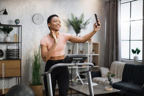 Attractive young sports african woman having online video call while working out, running, doing cardio training on treadmill in morning time, indoor on background of modern apartment or gym.
